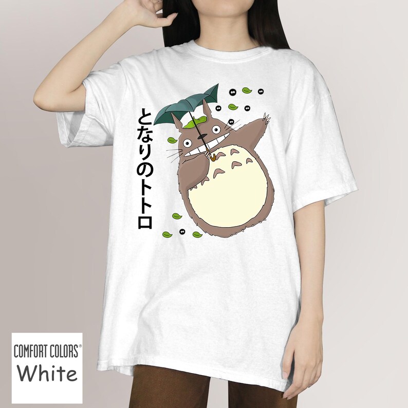 May include: White t-shirt with a cartoon Totoro character under an umbrella, with Japanese text and green leaf details. The shirt is labelled "Comfort Colors" and is a white, short-sleeved tee.