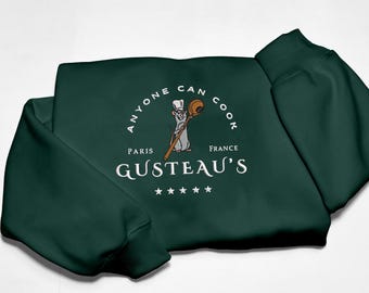 May include: Dark green sweatshirt with the text "ANYONE CAN COOK" in a circular design. The design includes a cartoon rat chef holding a wooden spoon. Below the text is "GUSTEAU'S" and "PARIS FRANCE".