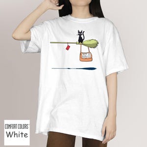May include: White t-shirt featuring a cartoon illustration of a black cat riding a broom. The broom has a red package and a bag that says "NOT IN SERVICE". The shirt is made of Comfort Colors material.