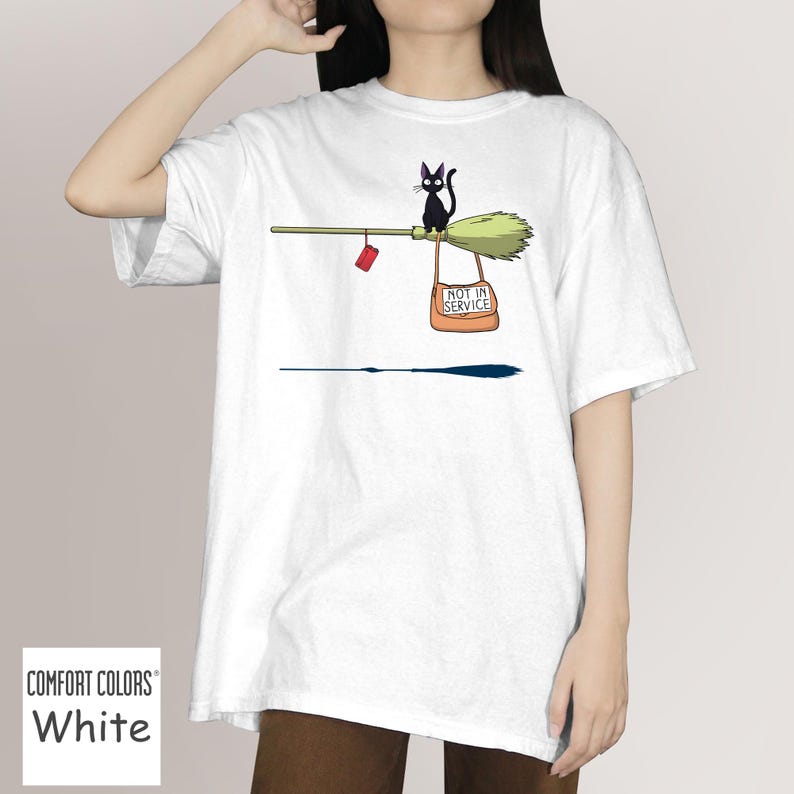 May include: White t-shirt featuring a cartoon illustration of a black cat riding a broom. The broom has a red package and a bag that says "NOT IN SERVICE". The shirt is made of Comfort Colors material.