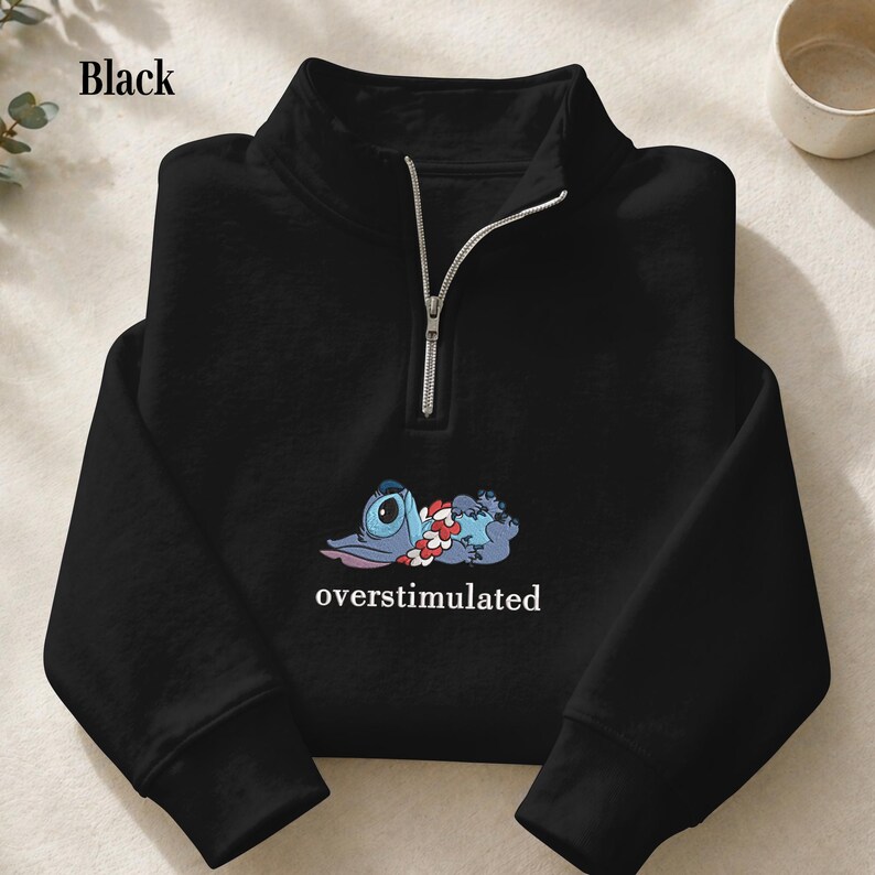 May include: A black quarter-zip sweatshirt featuring a graphic of a blue alien character wearing a floral lei, lying down. Below the character, the word overstimulated is printed in white text. The garment has long sleeves and a silver zipper.