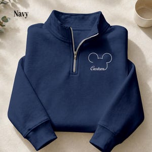 May include: Navy blue pullover sweatshirt with a quarter-zip closure and a white outline of a mouse head with the word "Custom" underneath. The sweatshirt has long sleeves and a mock neck. The word "Navy" is printed in the upper left corner.