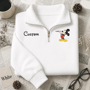 Puede incluir: Sudadera blanca con cuello con cremallera de un cuarto, con la palabra "Custom" en escritura negra y un gr&aacute;fico de Mickey Mouse. El personaje de dibujos animados sostiene un bol&iacute;grafo. La palabra "White" est&aacute; en una peque&ntilde;a tarjeta blanca.