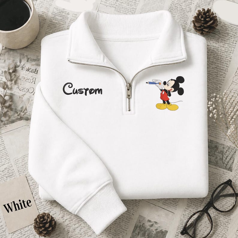 Puede incluir: Sudadera blanca con cuello con cremallera de un cuarto, con la palabra "Custom" en escritura negra y un gr&aacute;fico de Mickey Mouse. El personaje de dibujos animados sostiene un bol&iacute;grafo. La palabra "White" est&aacute; en una peque&ntilde;a tarjeta blanca.