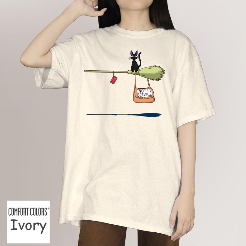 May include: Ivory Comfort Colors t-shirt featuring a whimsical illustration. A black cat sits on a broom with a sign that reads "NOT IN SERVICE." A small red object hangs from the broom. The shirt is a solid ivory color.
