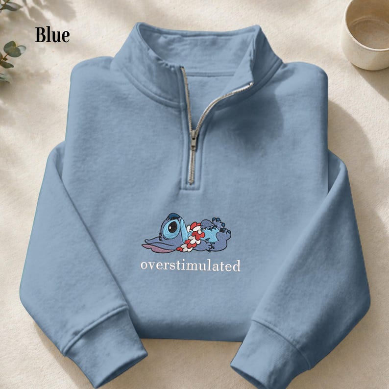 May include: A light blue quarter-zip sweatshirt featuring a graphic of a blue character wearing a red and white floral lei, lying down. Below the character, the word "overstimulated" is printed in white, lowercase, serif font.
