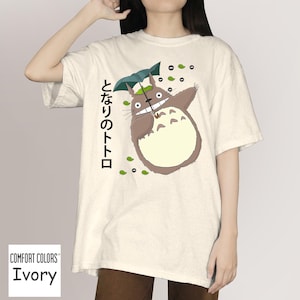 May include: Ivory Comfort Colors t-shirt with a cartoon Totoro design holding an umbrella. The design includes Japanese text and green leaf accents. The t-shirt has a relaxed fit.