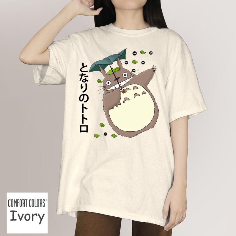 May include: Ivory Comfort Colors t-shirt with a cartoon Totoro design holding an umbrella. The design includes Japanese text and green leaf accents. The t-shirt has a relaxed fit.