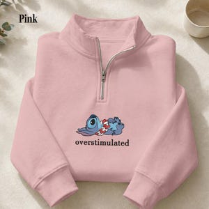 May include: A light pink quarter-zip sweatshirt featuring a graphic of a blue alien character wearing a floral lei, lying down with the text "overstimulated" printed in black underneath. The garment has a silver-toned metal zipper closure.