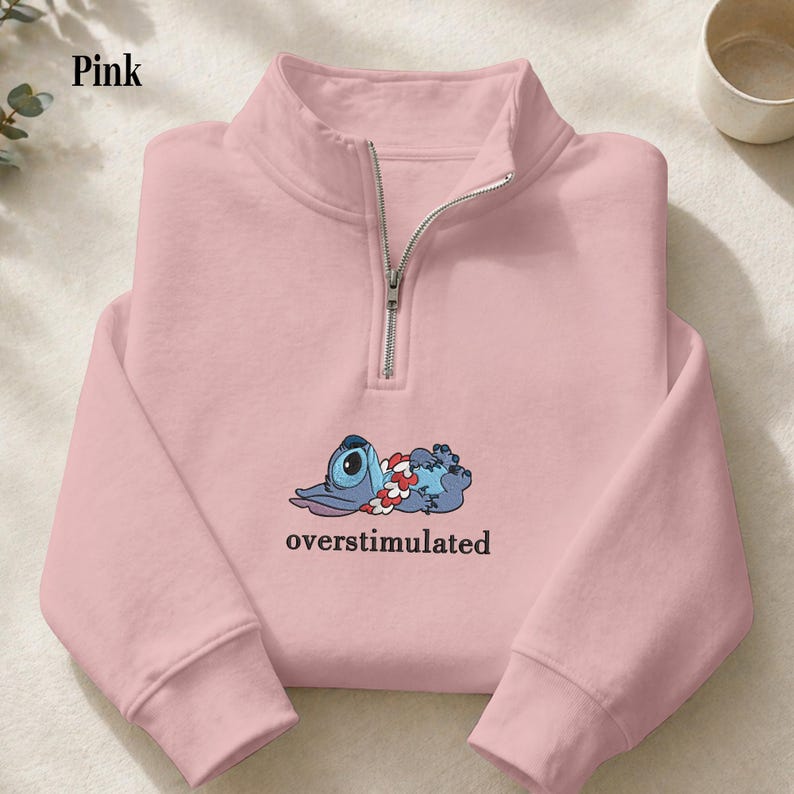 May include: A light pink quarter-zip sweatshirt featuring a graphic of a blue alien character wearing a floral lei, lying down with the text "overstimulated" printed in black underneath. The garment has a silver-toned metal zipper closure.