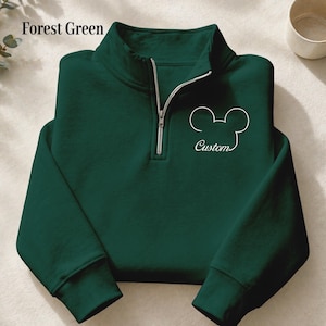 May include: Forest green quarter-zip sweatshirt with a white Mickey Mouse outline and the word "Custom" in script. The sweatshirt has a silver zipper and a high collar. The text "Forest Green" is in the upper left corner.