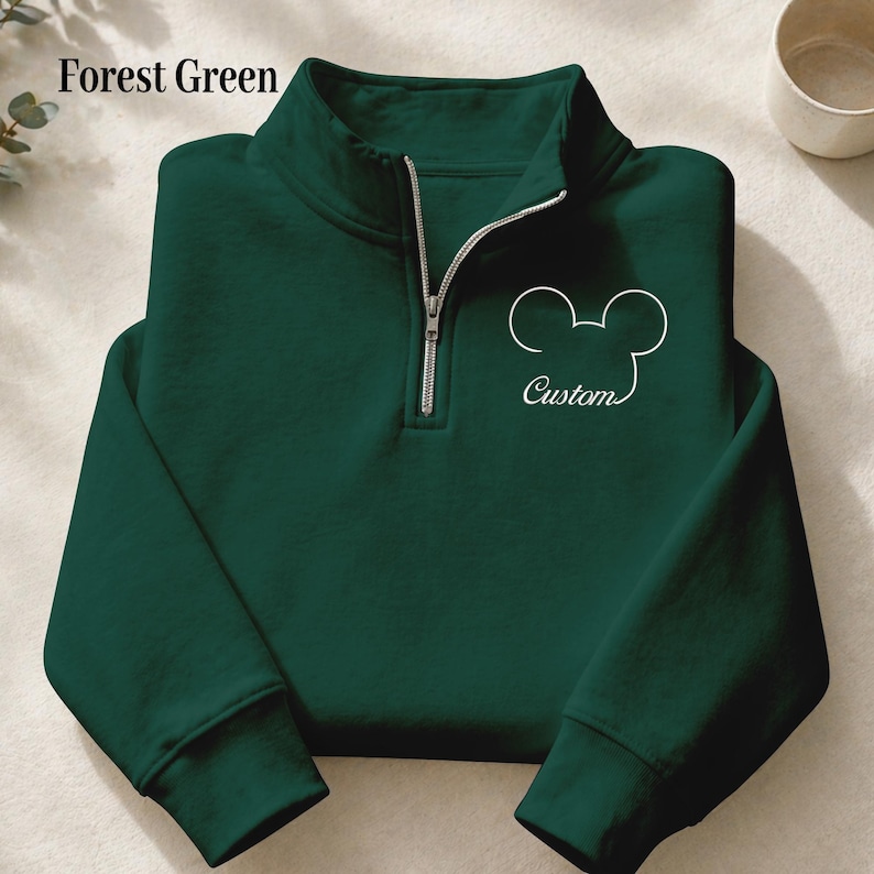 May include: Forest green quarter-zip sweatshirt with a white Mickey Mouse outline and the word "Custom" in script. The sweatshirt has a silver zipper and a high collar. The text "Forest Green" is in the upper left corner.