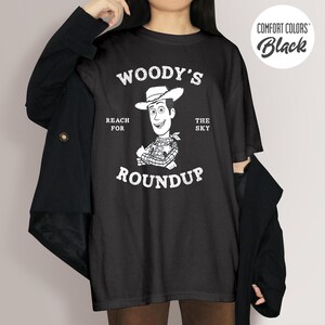 Pu&ograve; includere: T-shirt nero con una grafica di Woody di Toy Story. Il design include il testo "Woody's Roundup", "Reach for the Sky" e un'immagine a cartoni animati di Woody con un cappello da cowboy. La maglietta &egrave; etichettata "Comfort Colors Black".