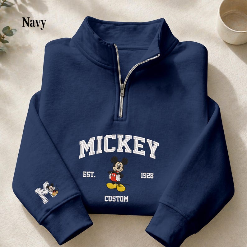 May include: A navy blue quarter-zip sweatshirt featuring a white collegiate-style graphic that reads MICKEY EST. 1928 CUSTOM with an illustration of Mickey Mouse. The left sleeve displays a large white letter M with a small Mickey Mouse graphic.