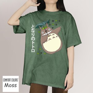 May include: Moss green t-shirt with a cartoon Totoro design, holding an umbrella, and Japanese text. The design includes leaves and soot sprites. The t-shirt has a relaxed fit and is a comfortable style.