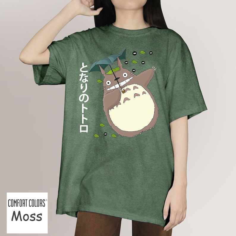 May include: Moss green t-shirt with a cartoon Totoro design, holding an umbrella, and Japanese text. The design includes leaves and soot sprites. The t-shirt has a relaxed fit and is a comfortable style.