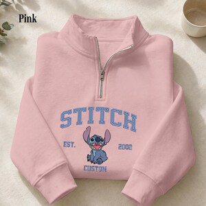 Pu&ograve; includere: Felpa rosa chiaro con collo con zip. La parte anteriore presenta la parola "STITCH" in lettere blu sopra un'illustrazione di un personaggio dei cartoni animati. Sotto, le parole "EST. 2002 CUSTOM".