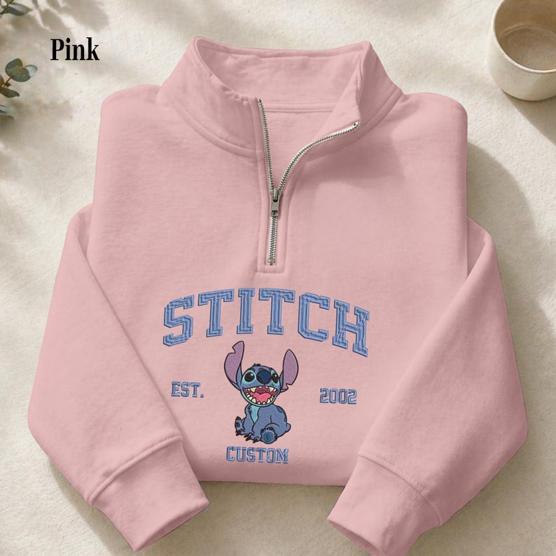 Pu&ograve; includere: Felpa rosa chiaro con collo con zip. La parte anteriore presenta la parola "STITCH" in lettere blu sopra un'illustrazione di un personaggio dei cartoni animati. Sotto, le parole "EST. 2002 CUSTOM".
