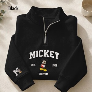 May include: A black quarter-zip sweatshirt featuring a white collegiate-style graphic that reads MICKEY EST. 1928 CUSTOM with an illustration of Mickey Mouse. The right sleeve displays a white letter M with a small Mickey Mouse graphic inside.