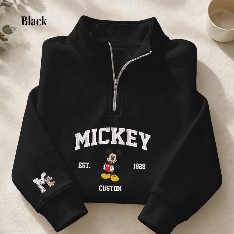 May include: A black quarter-zip sweatshirt featuring a white collegiate-style graphic that reads MICKEY EST. 1928 CUSTOM with an illustration of Mickey Mouse. The right sleeve displays a white letter M with a small Mickey Mouse graphic inside.