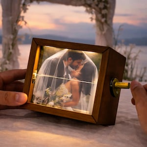 May include: A wooden music box featuring a photograph of a couple kissing. The box has a dark brown finish and a gold-coloured crank. The photograph is illuminated by a soft, warm light.