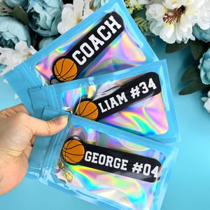 May include: Three iridescent blue zippered pouches with black and white basketball-themed keychains. The keychains have the names "COACH", "LIAM #34", and "GEORGE #04" printed on them.