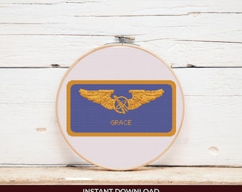 May include: A cross-stitch design in a wooden embroidery hoop. The design features a blue rectangle with gold wings and a symbol, and the name "GRACE" in gold. The hoop is set against a white wooden background.