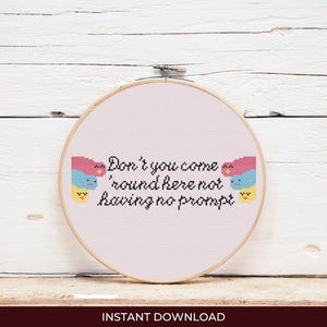 Puede incluir: Un bastidor de bordado de punto de cruz con el texto "Don't you come 'round here not having no prompt". El diseño incluye dos caras de dibujos animados con colores rosa, azul y amarillo. Las palabras están en hilo negro. El fondo es blanco.