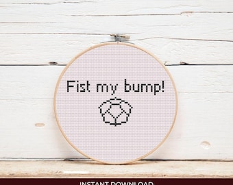 May include: A cross-stitch embroidery hoop with the text "Fist my bump!" in black, along with a black geometric design. The embroidery is set against a white fabric background, framed by a wooden hoop.