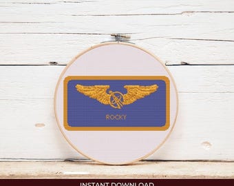 May include: Cross-stitch embroidery in a wooden hoop. The design features a blue rectangle with gold wings and a central emblem, and the name "ROCKY" in gold. The background is white, with the text "INSTANT DOWNLOAD" at the bottom.