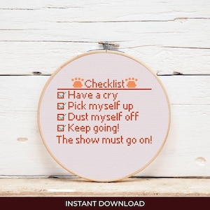 May include: A cross-stitch embroidery hoop with the text "Checklist" and a list of motivational phrases in orange thread. The phrases include "Have a cry," "Pick myself up," and "Keep going!" The background is a white wooden surface.
