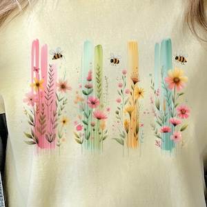 May include: A yellow t-shirt with a floral design and bees. The flowers are pink, yellow, and blue. The bees are black and yellow.