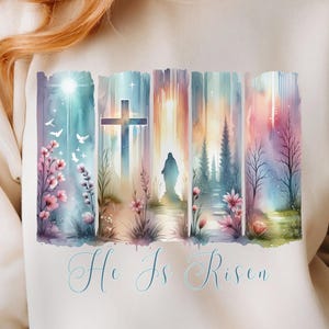 Könnte beinhalten: Ein beiger Pullover mit einem Aquarell-Design aus einem Kreuz, einer Figur, die davon weggeht, und Bäumen. Das Design ist von Blumen und Vögeln umgeben. Der Text "He Is Risen" ist unter dem Design gedruckt.