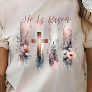 Könnte beinhalten: Ein weißes T-Shirt mit einem Aquarell-Design aus einem Kreuz und Blumen. Der Text "He Is Risen" ist in Rosa über dem Design geschrieben.