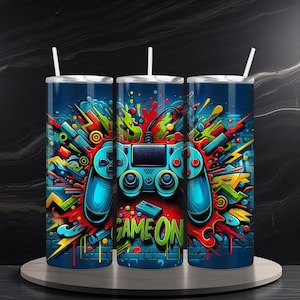 Puede incluir: Tres vasos de acero inoxidable con un diseño gráfico colorido que presenta un mando de videojuego y las palabras "Game On".