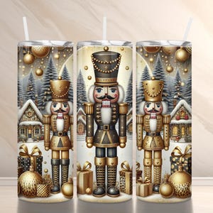 May include: Three stainless steel tumblers with a festive Christmas design. Each tumbler features a detailed illustration of a nutcracker soldier, surrounded by winter scenery, gifts, and ornaments. The design is predominantly gold, black, and white.