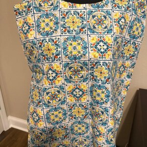 May include: A sleeveless top with a repeating tile pattern in yellow, blue, and white. The design features floral and geometric elements within each square, creating a vibrant and decorative look. The top is displayed on a black mannequin.