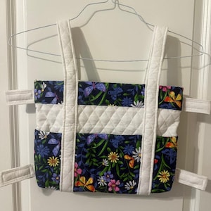 May include: A rectangular fabric bag featuring a dark blue background with a colorful print of butterflies, green leaves, and various flowers. The bag includes a central white quilted horizontal band, two white quilted shoulder straps, and four white side straps.