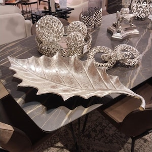 May include: A silver leaf-shaped serving tray with a textured surface. The tray is on a table with other decorative items, including silver orbs and a silver ring.