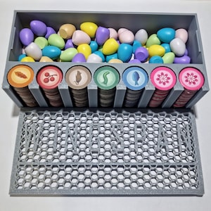 May include: A gray plastic organizer with a honeycomb pattern on the bottom and a compartment for storing game pieces. The organizer has eight compartments with colorful tokens and a top compartment with a collection of colorful plastic eggs.