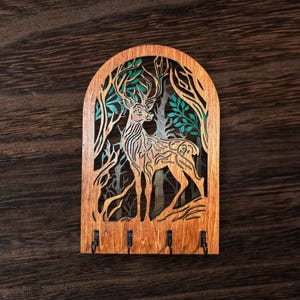 May include: Wooden key holder featuring a deer silhouette within a forest scene. The design is laser-cut with intricate details, including trees and foliage in shades of green and brown. Four metal hooks are positioned at the bottom for keys.