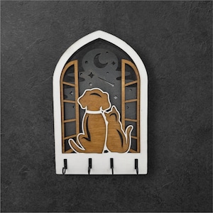 May include: A wooden key holder featuring a dog and cat silhouette looking out a window at a night sky with a crescent moon and stars. The key holder has a white frame and four black hooks.
