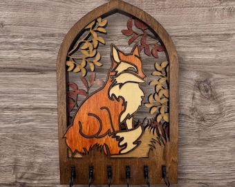 May include: Wooden key holder featuring a fox design. The fox is depicted in shades of orange and cream, set against a backdrop of leaves and branches. The key holder has five black hooks and a dark brown frame.