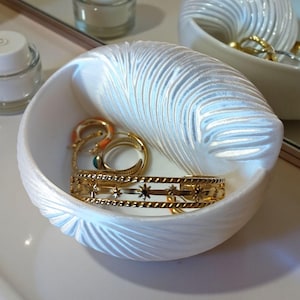 May include: White, shell-shaped jewelry dish with a textured, ridged design. The dish holds gold rings and a gold bracelet with star cutouts. A second dish with jewelry is visible in the background.