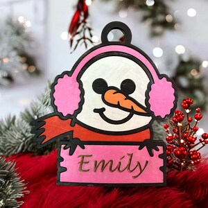 May include: A wooden snowman ornament with pink earmuffs and a carrot nose. The snowman is wearing a red scarf and holding a pink sign that reads "Emily" in gold lettering. The ornament has a black outline.