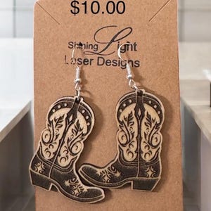 May include: A pair of wooden cowboy boot earrings with a brown wood finish and a black and white design. The earrings are on a card that says "Shining Light Laser Designs" and "$10.00".