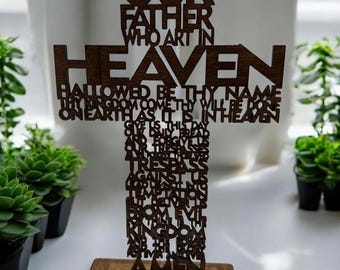 May include: A wooden cross with the text "OUR FATHER WHO ART IN HEAVEN" and the Lord's Prayer. The cross is brown and stands on a rectangular wooden base. The text is cut out of the wood.