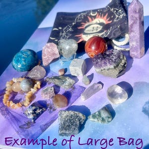 May include: An assortment of crystals and gemstones, including a blue sphere, rose quartz, amethyst, and a red and white orb. A black bag with a sun and moon design is visible. The text "Example of Large Bag" is at the bottom.