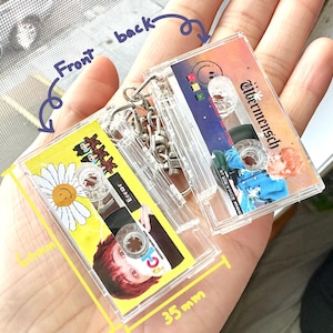 May include: Two clear cassette tape keyrings, 3.5cm x 6cm, held in a hand. One keyring has a yellow background with a daisy and the word "ERROR". The other has a colourful design and the word "Albermensch". Both have silver keyrings.