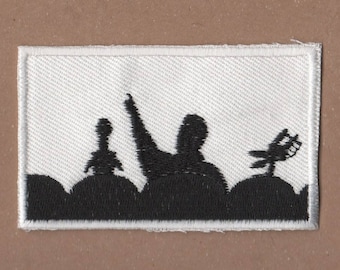 May include: Embroidered patch featuring a black silhouette of a person with an arm raised, a microphone, and a satellite dish. The patch has a white background and a silver border, suitable for jackets or bags.